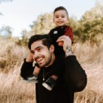 Man with a black sweater on outside with his 1 year old son on his shoulders smiling with hay/grass in the background